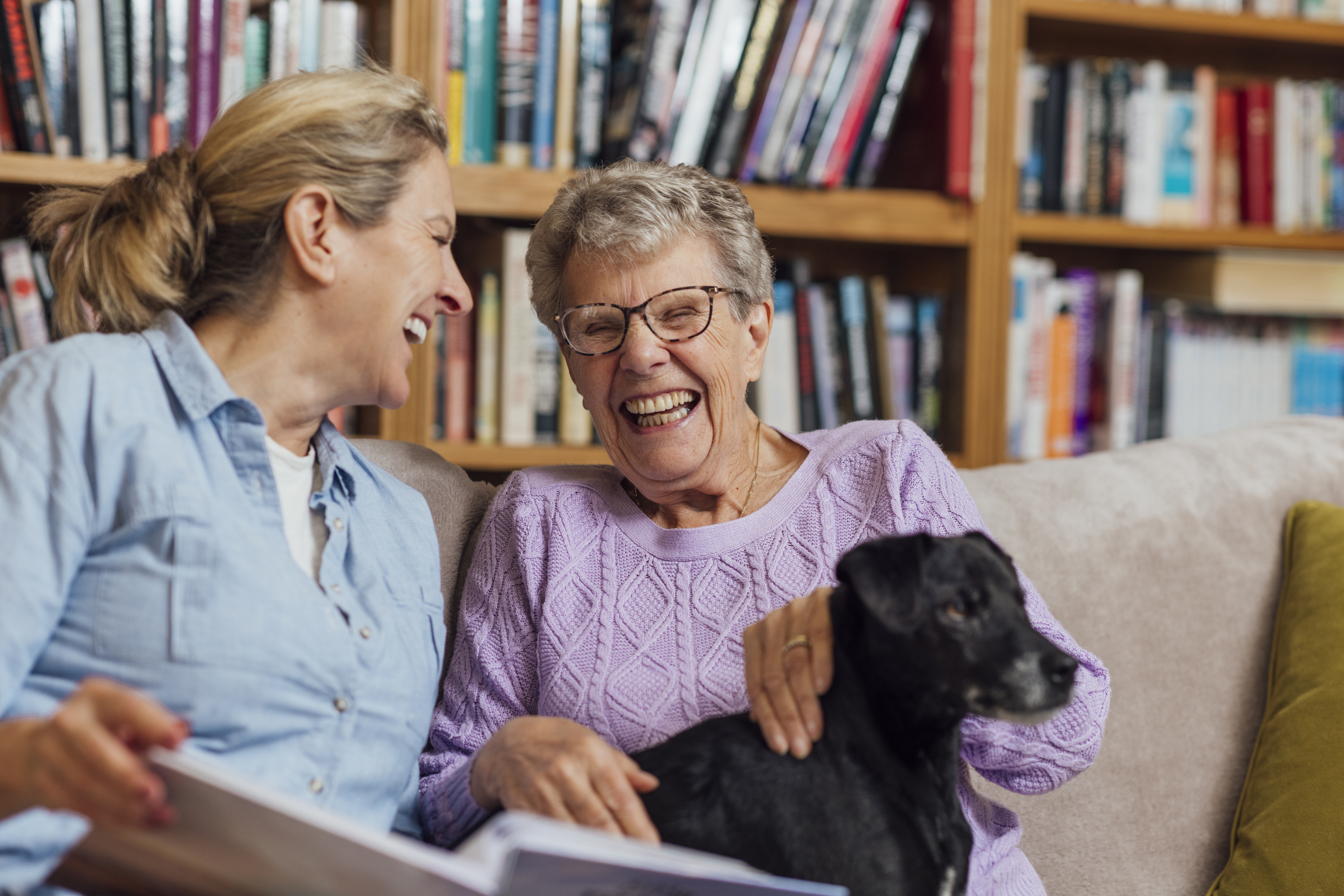 Image of an elderly mother and her daughter, smiling whilst reading a book on the sofa.