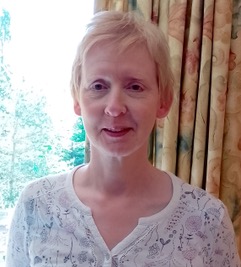 A portrait image of Alison. She is stood in front of a window, wearing a white floral top, smiling.
